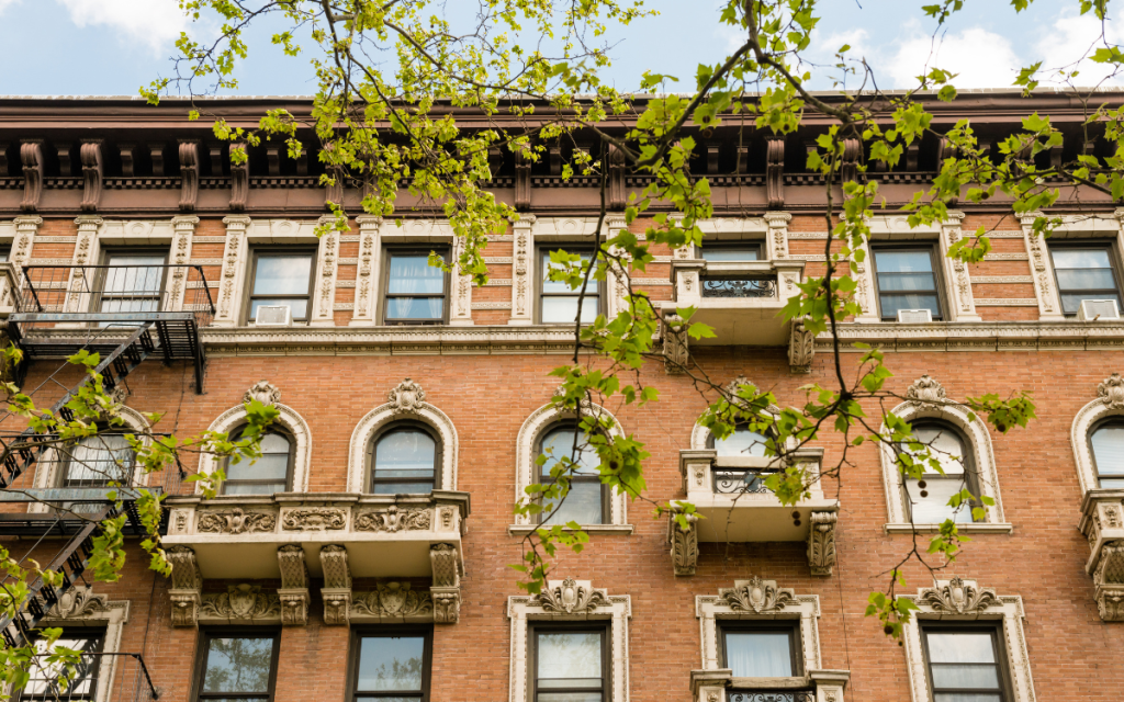 Image of a pre-war, brick facade walk up building in NYC with elaborate window surrounds and lintels. The brick is a light orange brown. Newly greened branches from a tree are in the foreground. 