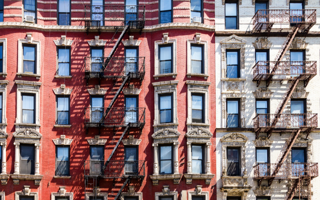 Image of brick facade pre-war walk-up style buildings in NYC. The buildings to the left are red brick while the buildings to the right are white brick. both have elaborate lintels and window surrounds. 